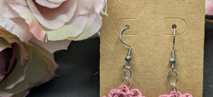 a pair of pink crocheted earrings in the shape of a flower against a black backdrop with silk flowers