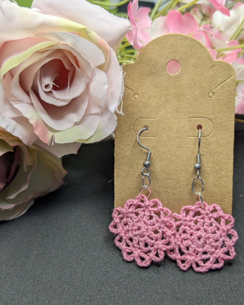 a pair of pink crocheted earrings in the shape of a flower against a black backdrop with silk flowers